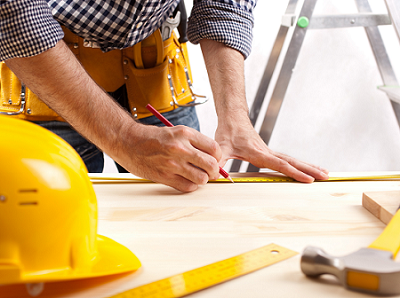 Contractor with sleeves rolled up stands over work table measuring some wood there is an open ladder next to him and a yellow hard hat and hammer on the table in front of him